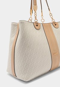 Sac fourre-tout en matière beige texturée avec un motif géométrique. Comprend des accents lisses couleur camel, une ferrure dorée, et des doubles anses.