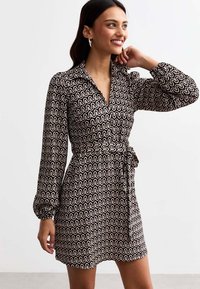 Robe portefeuille noire et blanche avec un motif ondulé, manches longues, décolleté en V et une ceinture à nouer à la taille, fabriquée en tissu doux avec un léger éclat.