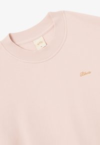 Sweatshirt rose clair avec col côtelé et texture douce. Présente une broderie dorée subtile du nom de la marque sur le devant. Taille XL.