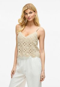 Top de tanque de ganchillo beige con diseños circulares y dobladillo en forma de concha, combinado con pantalones blancos de corte holgado. Tirantes de espagueti, textura ligera.