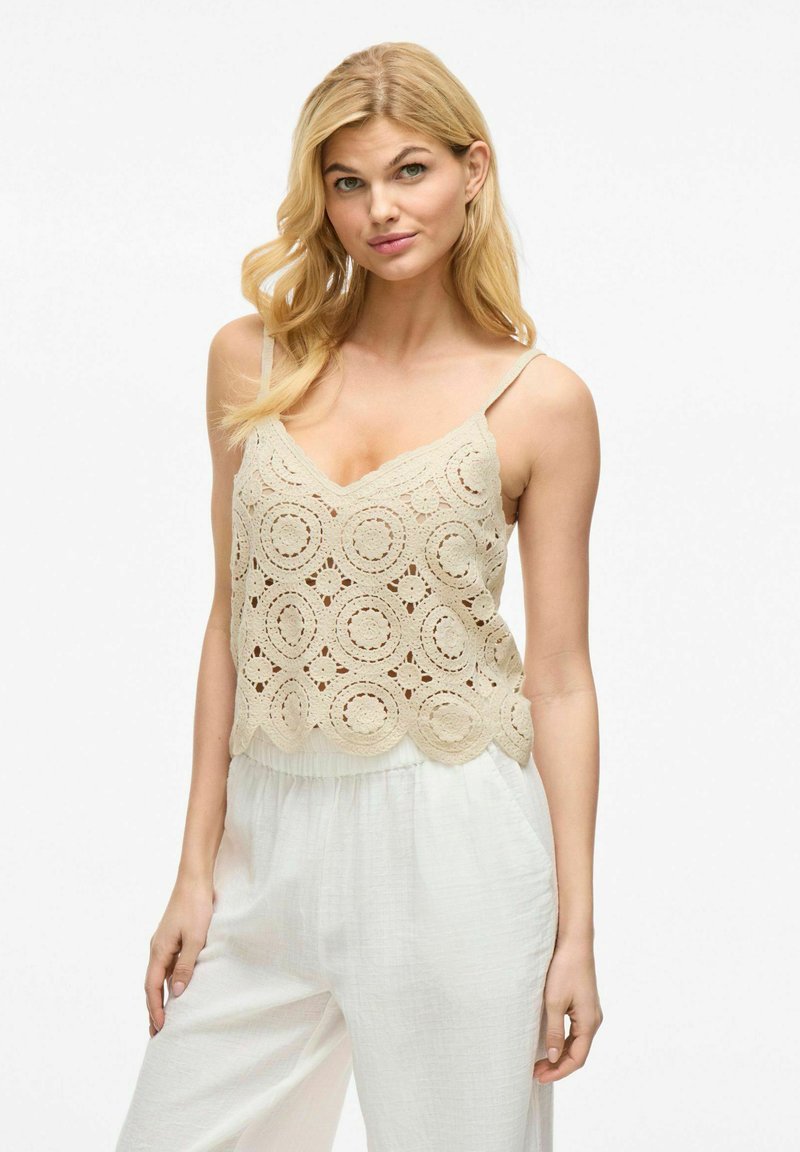 Top de tanque de ganchillo beige con diseños circulares y dobladillo en forma de concha, combinado con pantalones blancos de corte holgado. Tirantes de espagueti, textura ligera.