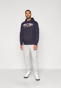 Mørkeblå hættejakke med "NEW YORK" grafik, kengurulomme og snore, kombineret med grå cargo sweatpants og hvide sneakers.
