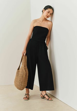 Frau in schwarzem trägerlosen Jumpsuit und Sandalen, die eine große hellbraune geflochtene Tasche hält und an einer weißen Eckwand lehnt.