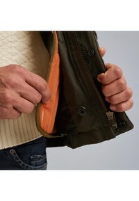 Maslinasto zelena jakna s narančastim unutrašnjim dijelom, sa zatvaračem i kopčanjem na dugmad. Pod njom se vidi pletena vunena majica.