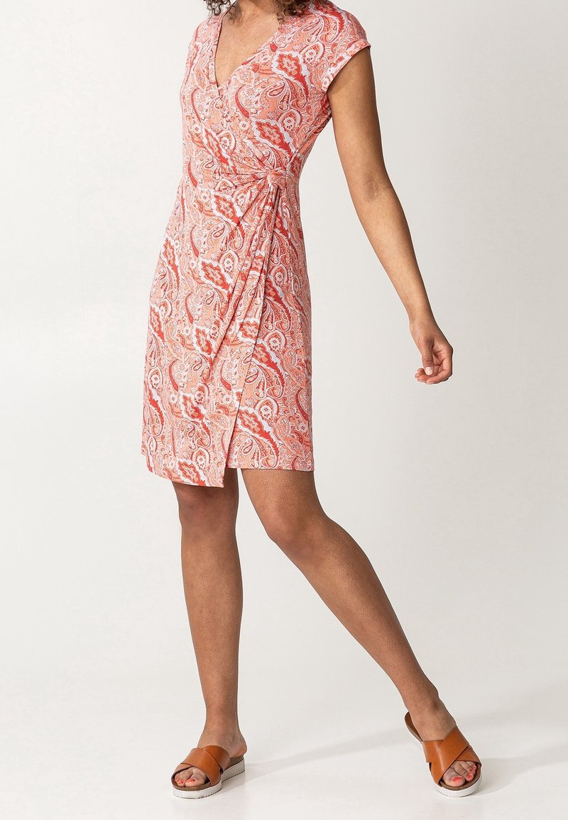 Sukienka o krótkim rękawie, zapinana na zakładkę w kolorze koralowym z wzorem paisley. Posiada dekolt w kształcie litery V, ściągniętą talię oraz długość do kolan. Noszona z brązowymi klapkami.