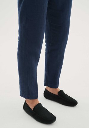 Piernas con pantalones ajustados azul oscuro y mocasines negros sin cordones sobre un fondo claro, mostrando un estilo de calzado informal.