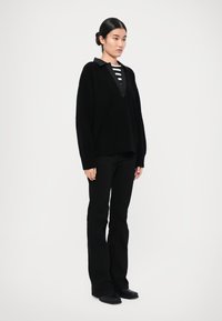 Schwarzer übergroßer Strickpullover mit V-Ausschnitt, kombiniert mit einem schwarz-weißen gestreiften Oberteil, schwarzen ausgestellten Hosen und flachen schwarzen Schuhen.