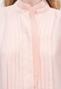 Κοντινή λήψη ενός ανοιχτόχρωμου ροζ μπλουζιού με κάθετες πιέτες και μικρό στρογγυλό γιακά που στερεώνεται με ένα κουμπί.