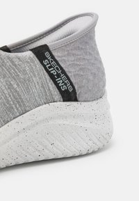 Gros plan sur le talon d'une chaussure Skechers slip-on grise avec semelle blanche tachetée et étiquette en tissu sur le côté.
