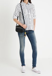 Camisa de botones a rayas blancas y negras, jeans desgastados azules, bolso cruzado negro con borlas y zapatillas blancas.