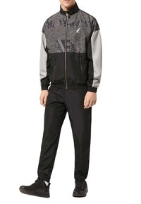 Tuta sportiva con giacca a zip nera e grigia a motivi, maniche lunghe, abbinata a pantaloni neri coordinati e sneakers nere.