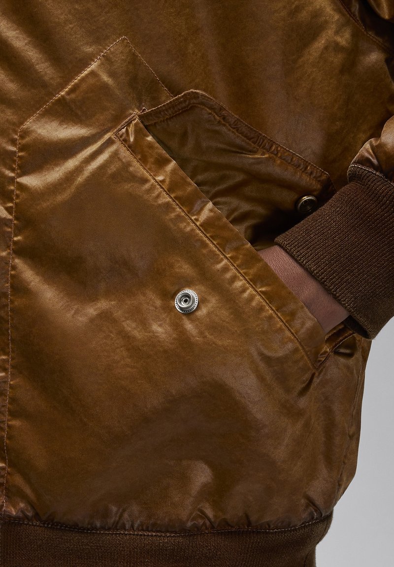 Braune Leder-Bomberjacke mit metallischem Druckknopfverschluss an der Tasche, gerippten Bündchen und einer glatten Textur. Verfügt über ein diamantförmiges Taschendesign.