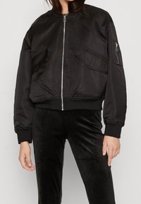 Blouson aviateur noir à finition satinée, fermeture zippée à l'avant, deux poches avant, et poignets ainsi qu'ourlet côtelés. Associé à un pantalon en velours noir.