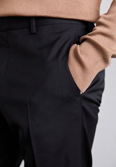 Μαύρο tailored παντελόνι από απαλό ύφασμα, με πλαϊνές τσέπες και στενή εφαρμογή, σε συνδυασμό με έναν ανοιχτό καφέ υφασμάτινο πουλόβερ.