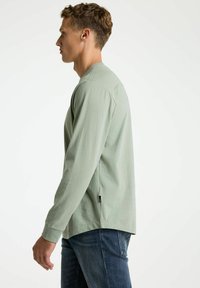 Cămașă cu mânecă lungă dintr-un material verde moale, croi lejer, decolteu rotund, slituri laterale și detalii cu etichetă vizibile la margine. Asortată cu blugi din denim.
