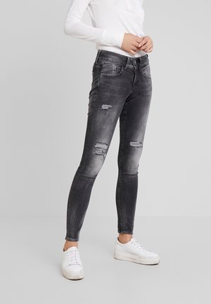 Kvinde iført falmede sorte, slidte skinny jeans, hvid langærmet top og hvide sneakers, stående på en neutral grå baggrund.