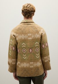 Giacca di lana in tonalità nocciola con motivi geometrici in crema, verde e bordeaux. Presenta un colletto a risvolto e maniche di lunghezza media. Superficie testurizzata.