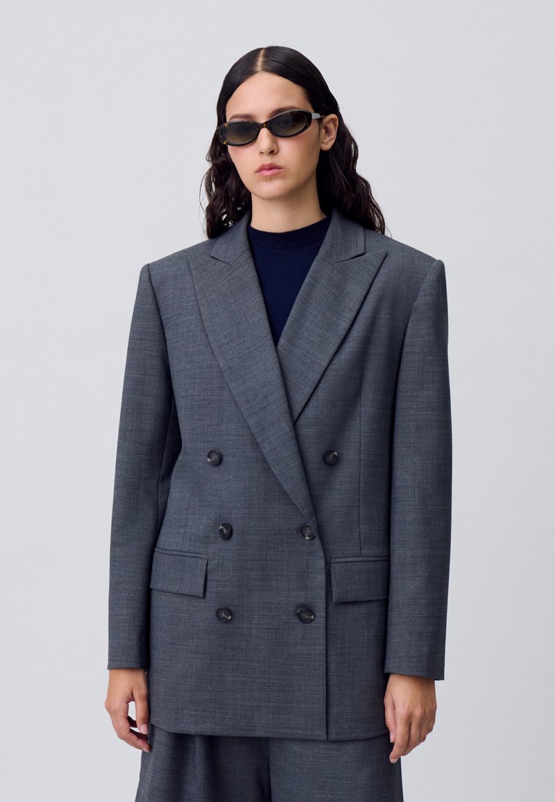 Blazer grigio doppiopetto con ampi risvolti, due tasche frontali e un tessuto texturizzato. Indossato sopra un top scuro, abbinato a occhiali da sole eleganti.