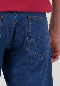 Donkerblauwe denim jeans met een achterzak, subtiele stikseldetails en een gladde textuur. Draag met een gedeeltelijk zichtbare rode top.