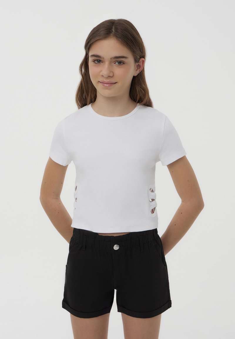 T-shirt bianca a coste con maniche corte e dettagli tagliati ai lati, abbinata a pantaloni corti neri con bottone e vita elasticizzata.