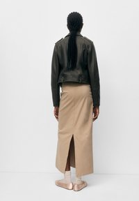 Kvinna med flätat hår bär en svart läderjacka, beige lång kjol med slits bak, vita strumpor och rosa ballerinas, stående med ansiktet mot väggen.