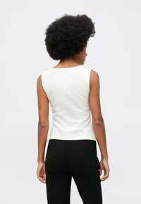 Kobieta z fryzurą afro, ubrana w białą bluzkę bez rękawów i czarne spodnie, pokazana plecami na tle białego, gładkiego tła.