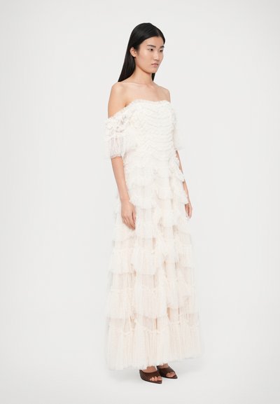 Off-the-shoulder, layered gown in soft cream, featuring ruffled tulle, sheer sleeves, and textured fabric with dotted patterns. Flats worn.