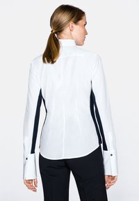 Camicia bianca con pannelli laterali blu navy, maniche lunghe e dettagli dei bottoni neri. Tessuto liscio e vestibilità sartoriale, con un design sul retro.
