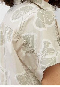 Lichtbeige shirt met een groot, gedempt groen bloemenpatroon. Heeft korte mouwen, een klassieke kraag en plooien langs de naad voor een relaxte pasvorm.