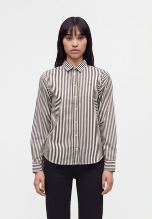 Femme aux cheveux noirs et lisses portant une chemise à rayures verticales marron et blanche à manches longues boutonnée et un pantalon noir, debout devant un fond uni.