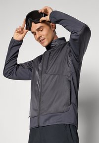 Siva zip-up jakna z visokim ovratnikom, iz lahkega blaga, stranskimi žepi in kontrastno teksturo. Model prilagaja črne smučarske očala.