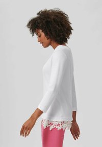 Witte longsleeve top met een ronde halslijn, voorzien van een bloemenkant zoom. Gecombineerd met roze slim-fit broek, die een slanke silhouet laat zien.