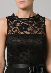 Haut en dentelle noire avec un col haut, présentant des motifs floraux et une jupe noire lisse avec une ceinture en satin. Design structuré et ajusté.