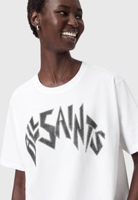 T-shirt bianco in cotone con una grafica audace in strass neri che riporta la scritta "SAINTS". Maniche corte e scollatura rotonda. Texture liscia.