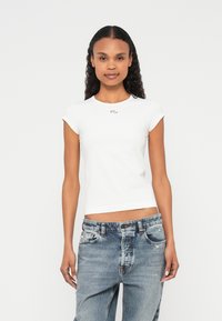 Balta pieguloša T-krekla ar cepures piedurknēm, apaļu kakla izgriezumu un mazu logotipu krūtīs, kopā ar gaiši zilām bojātām džinsa biksēm.