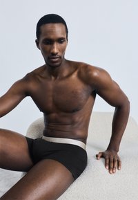 Fekete boxeralsó világosszürke derékpánttal, amelyen szöveg található. A modell izmos testalkatú, és egy texturált krémszínű felületen ül.