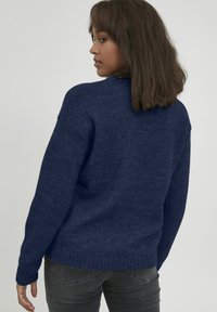 Dunkelblauer Pullover mit strukturierter Oberfläche, hoher Kragen, langen Ärmeln und geripptem Saum. Kombiniert mit dunkelgrauen Jeans.