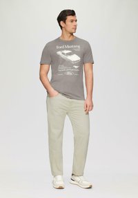 Szary T-shirt z grafiką Ford Mustang, zestawiony z jasnobeżowymi spodniami i białymi sneakersami z czarnymi akcentami.