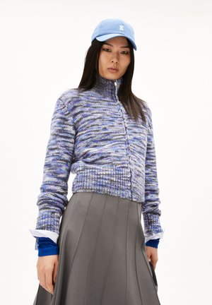 Gilet tricoté en bleu et violet avec un col montant, associé à une jupe plissée grise. Dispose de poignets côtelés et d'une fermeture à boutons sur le devant.