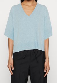 Vaaleansininen, lyhythihainen v-pääntie-neuletakki rentolla istuvuudella, pehmeällä tekstuurilla ja lyhyt helma, yhdistettynä mustiin narulla kiristettäviin housuihin.