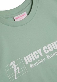 Šviesiai žalia trumpų rankovių medvilninė marškinėlių su balta grafika ir tekstu "JUICY COUTURE", vaizduojančiu figūrą judesyje, su apvalia iškirpte.