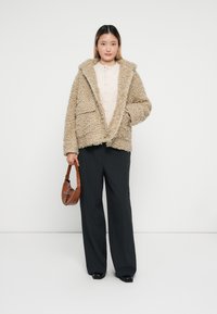 Giacca beige soffice con cappuccio e due tasche frontali, indossata sopra un maglione chiaro e abbinata a pantaloni neri a righe sottili e una borsa marrone.