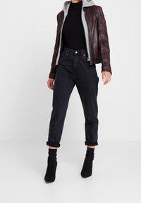 Person klädd i svarta högmidjade uppvikta jeans, svarta sockboots, svart polotröja, grå huvtröja och brunt läderjacka som står på ett vitt golv.