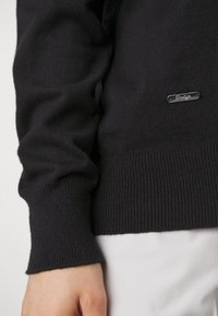 Svart stickad tröja med långa ärmar, ribbade manschetter och en liten silverfärgad logotagg framtill. Mjuk textur, kroppsnära design.