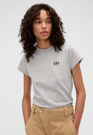 Kobieta o kręconych włosach nosi jasnoszary t-shirt GAP i beżowe spodnie z wysokim stanem, stoi swobodnie z rękami w kieszeniach.