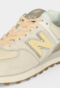 Μπεζ αθλητικό παπούτσι New Balance 574 με ανοιχτοκίτρινες κορδόνια, κίτρινο λογότυπο "N", ύφασμα από δίχτυ και λευκό επενδεδυμένο γιακά.