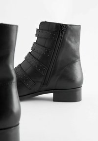 Czarne skórzane botki do kostki z zamkiem błyskawicznym, z szerokimi paskami z ćwiekami i niskim słupkowym obcasem. Gładka tekstura i dopasowany krój.