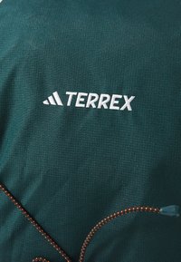 Zeleni tkanina s teksturom i bijelim logotipom "TERREX". Ima narančaste detalje na vezici. Prikazuje izblizak uzorka tkanine.