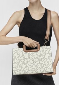 Beżowa torebka z teksturowanym białym wzorem, brązowymi uchwytami i odczepianym paskiem. Projekt cechuje się zakrzywionymi liniami i uporządkowanym kształtem.