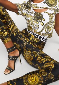 Schwarze Leggings mit einem goldenen, aufwendigen Muster, kombiniert mit einem weißen Oberteil mit ähnlichen goldenen Akzenten und Cut-Out-Details. Schwarze, mit Nieten verzierte Riemchensandalen.